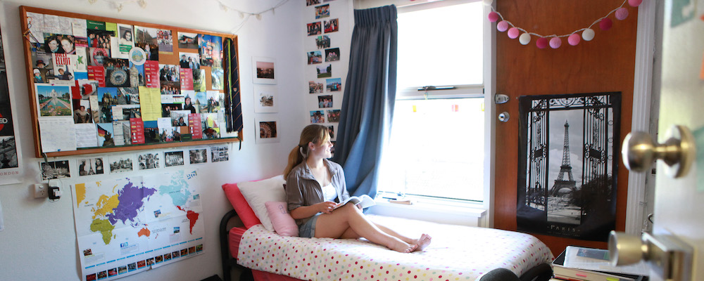 A young woman sits on her bed, looking out the window, in a brightly decorated room in student
    accommodation.