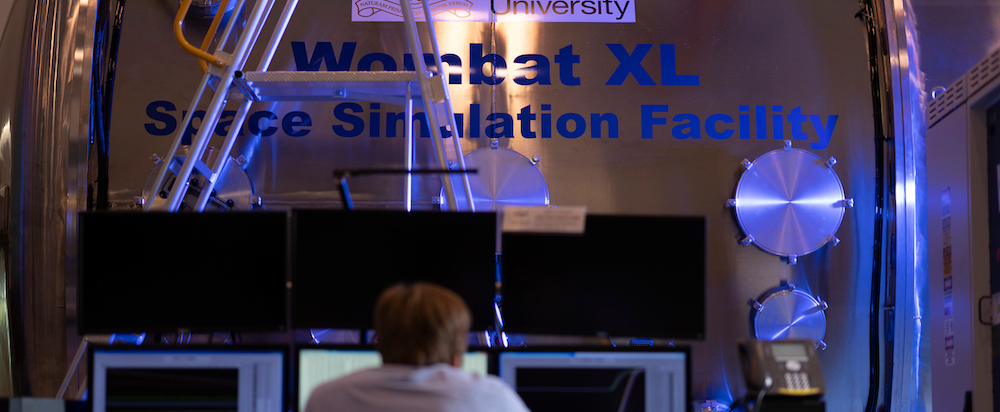 A man sits at an array of monitors, with the Wombat XL behind his desk.