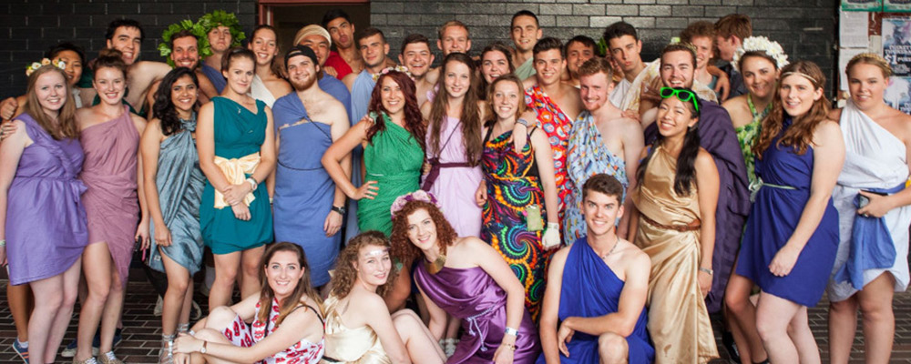 A large group of students in colourful togas pose in front of the entrance to Griffin Hall.