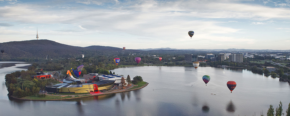 Hot air balloons hover above Lake Burley Griffin with ANU in the distance.