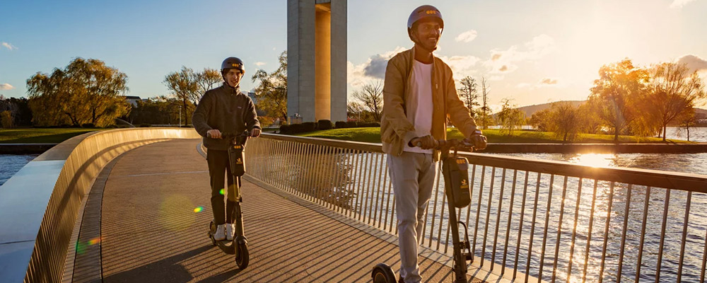 Two men ride electric scooters across the bridge from the Carillion on Lake Burley Griffin.
