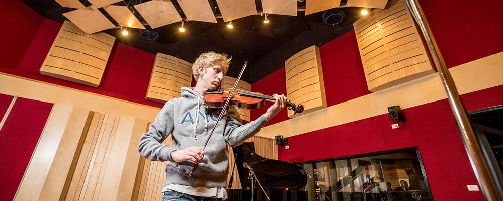 An ANU student plays violin in a recording studio, as other students watch from the mixing desk.