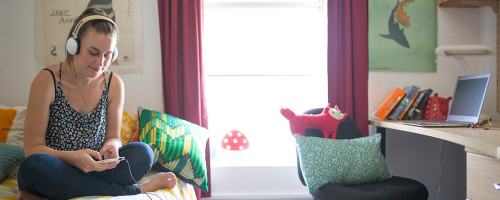 A woman sitting on her bed and listening to music in her student accommodation room.