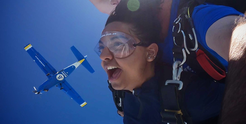  Priya and a skydiving instructor fall through the air, high up in the sky. Priya is screaming.