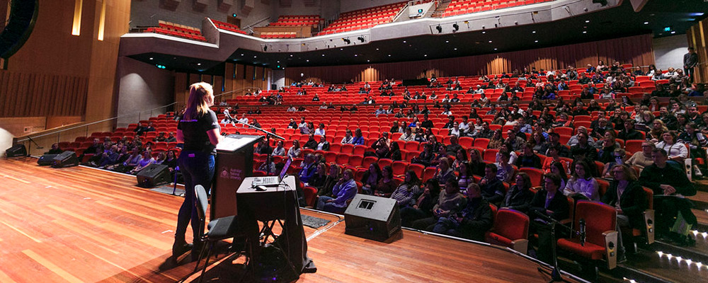 A woman stands at a lectern and delivers a presentation to a large group of ANU students in a theatre.