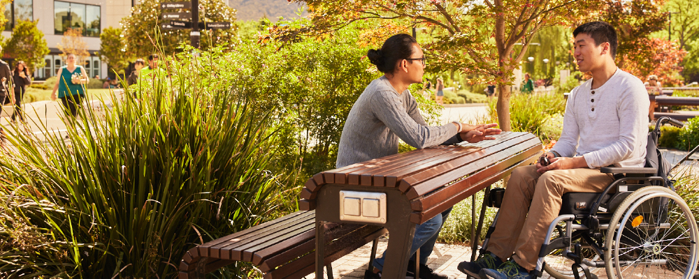 Two students chat at an outdoor bench on a sunny day.