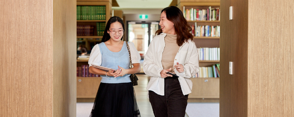 Two ANU students walking through a corridor with books visible behind them.