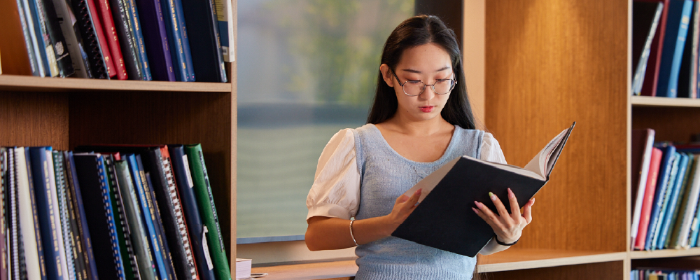 An ANU student in a small library reading a large book.