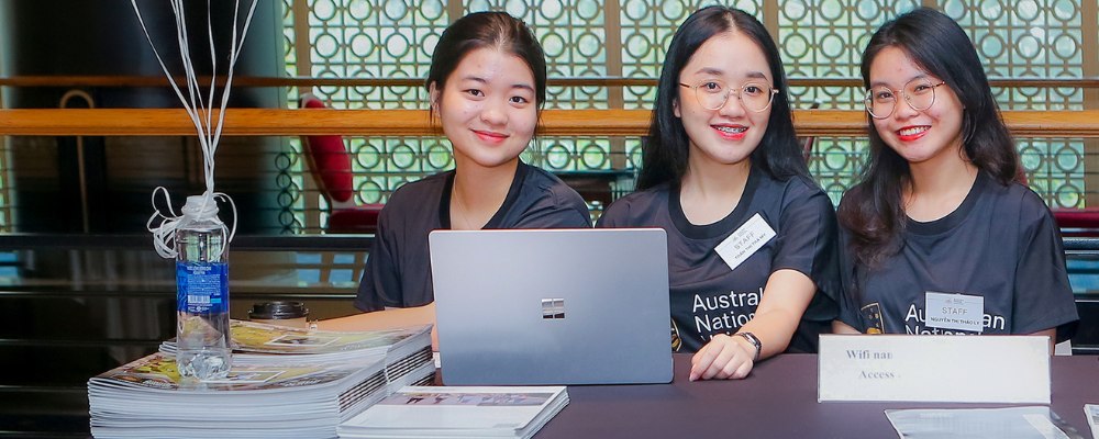 Three women sit at an ANU Information Day help desk in front of a laptop.