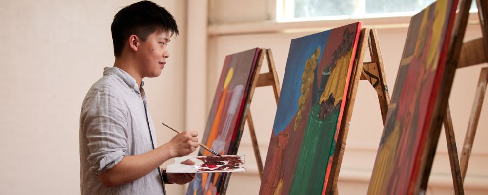 ANU student standing in front of paintings on easels, holding a palette in one hand and a paint brush in another.