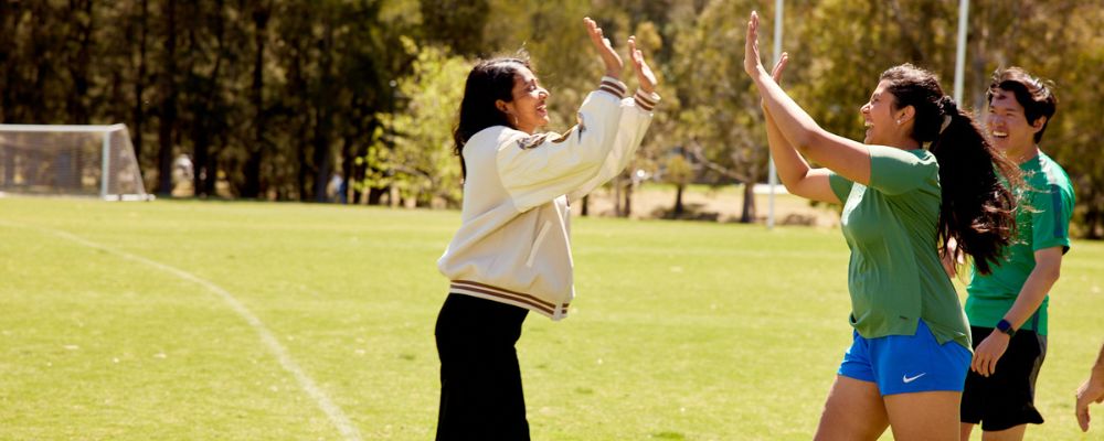ANU students high-fiving and celebrating on an ANU campus oval, playing football.