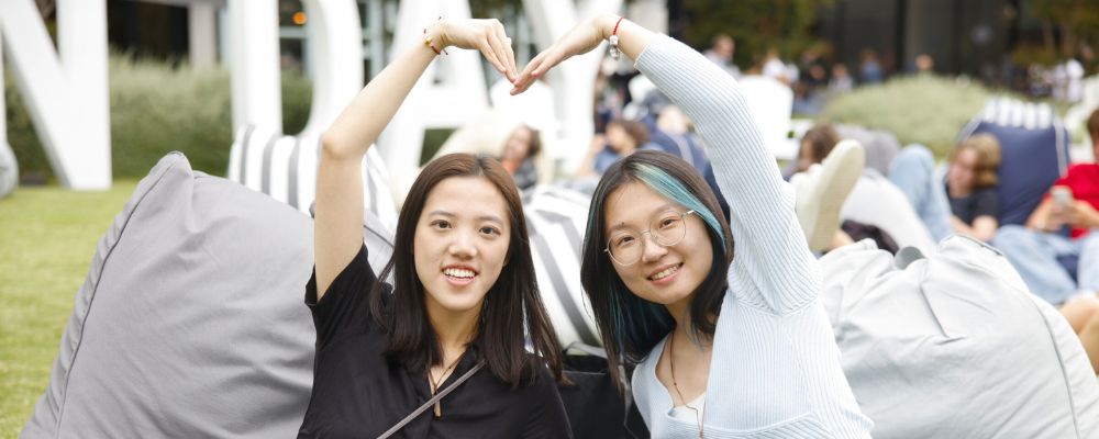 Two young women attending ANU Open Day sitting on Kambri Lawns making a heart with their bodies and smiling at the camera.