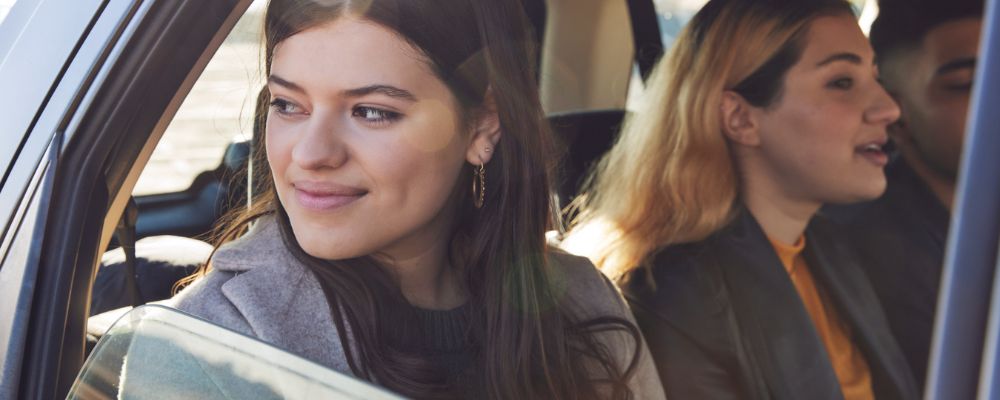 ANU student looks out the window of a car with a smile on her face, two ANU students in the background, also sitting in the backseat of the car.