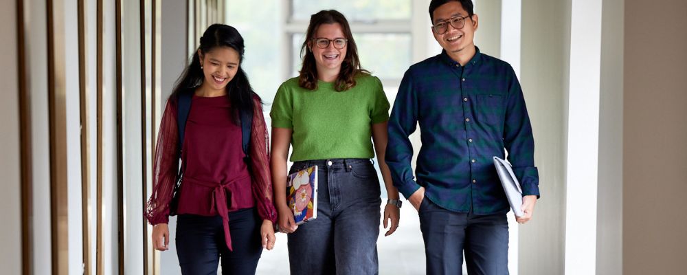 Three ANU students walk down a hall with books in hand, smiling.