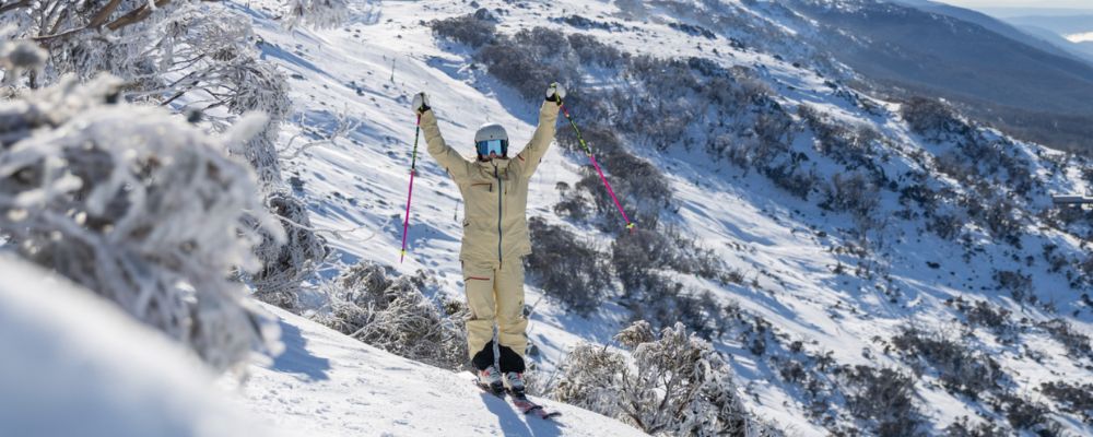 A woman in full ski gear stands on the edge of a snow mountain and poses for the camera in Thredbo.