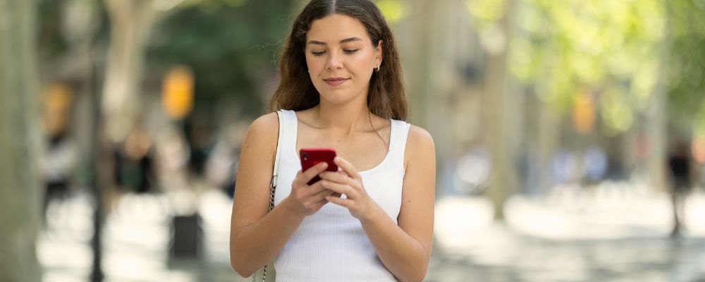 Female student scrolls on mobile device as she walks along an outside street.