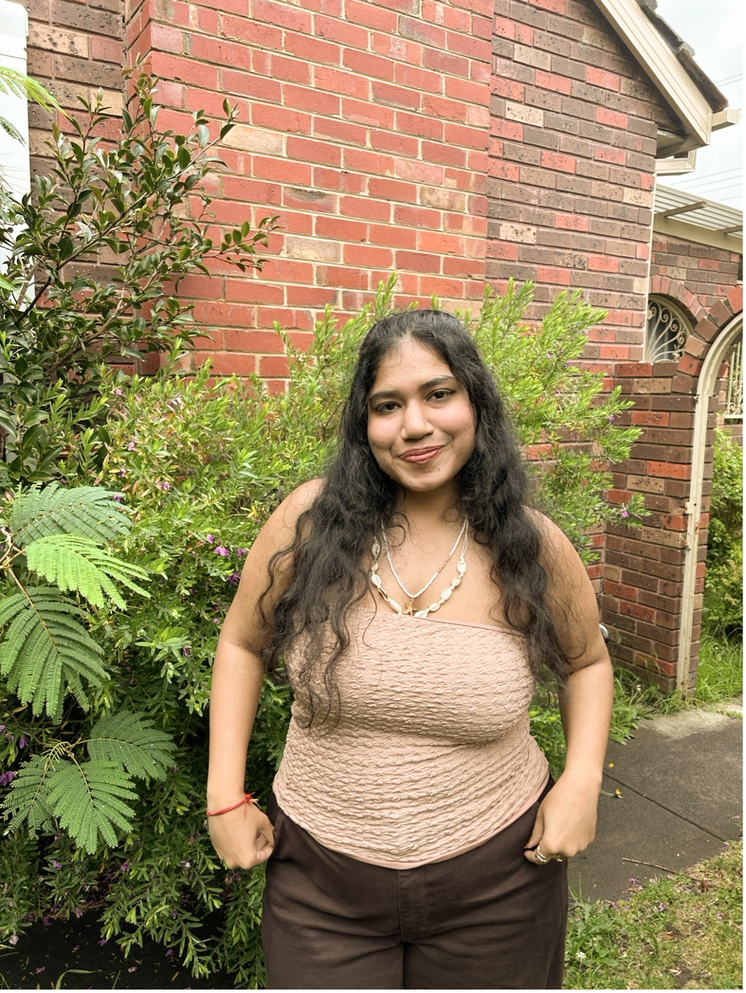 Disha stands outside a house in Canberra with greenery behind her, hands on her hips.