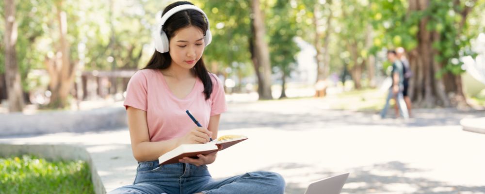 A female postgraduate student sits outside, studying and listening to a podcast on her device.