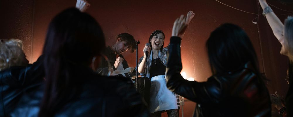A band with a female lead performer stands on a stage and performs in front of a live crowd.