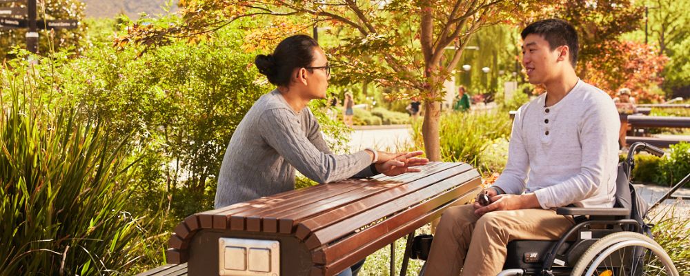 Two friends at ANU sit together at one of the outside seating benches in the Kambri precinct, talking and laughing.
