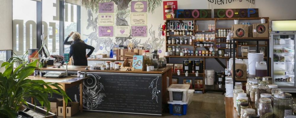 The Food Co-Op store interior in Canberra with shelves of pantry goods, jars, plants and a service counter near large front windows.