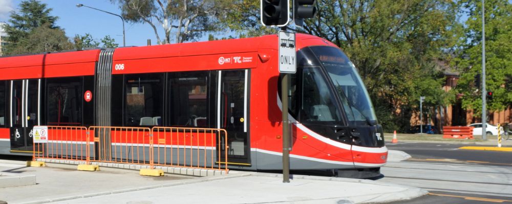 The Canberra light rail stopped at a station on a sunny day, with trees and a road intersection in the background.