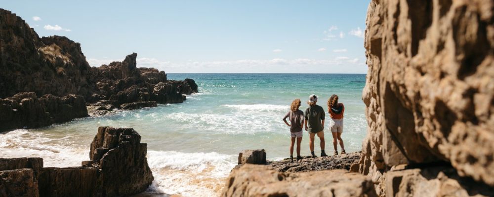 Three people stand on a rocky coastline looking out at the ocean, with waves breaking between cliffs.