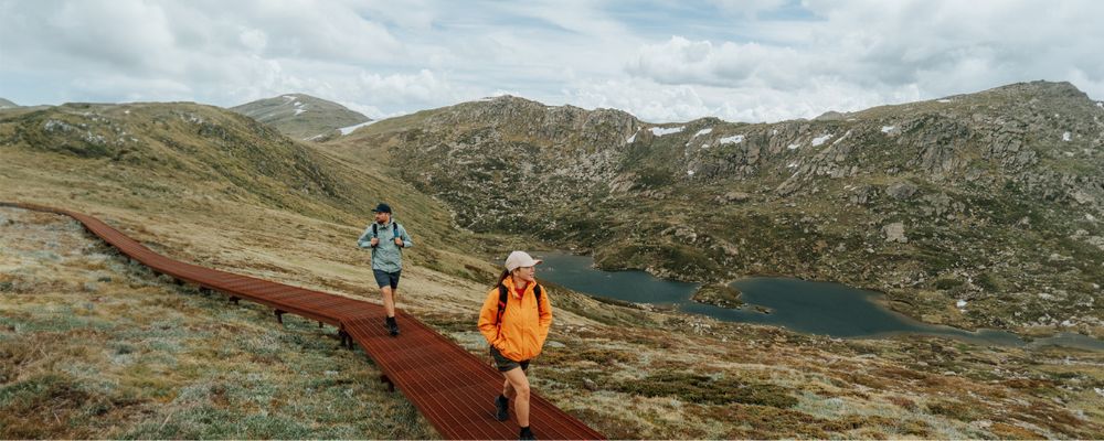 Two hikers walk along a raised track through the Snowy Mountains, with rocky hills and alpine lakes in the background.