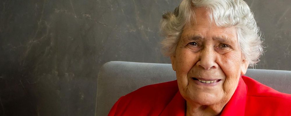 Lowitja O’Donoghue smiles for a portrait, seated against a dark background and wearing a red jacket.
