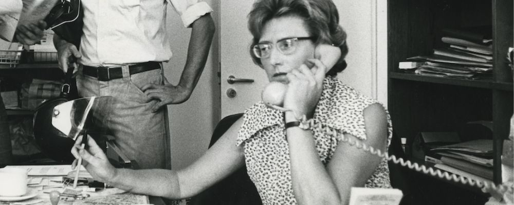 Di Riddell sits at a desk at ANU, holding a telephone receiver while working with papers and office materials around her.