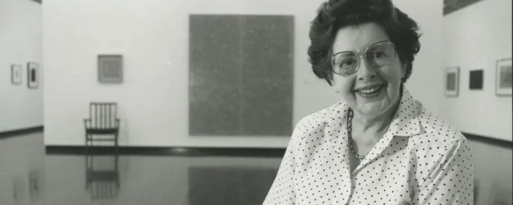 Pauline Griffin smiles for a black and white portrait inside Drill Hall Gallery, with framed artworks and a chair behind her.