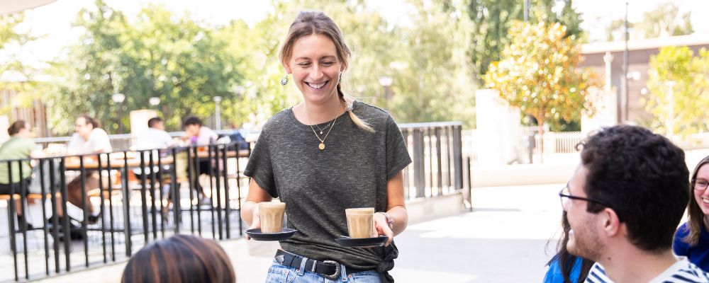 A worker at ANU carries two coffees while smiling and speaking with a group seated at an outdoor campus café