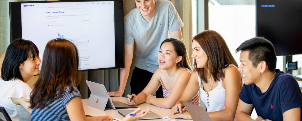 A group of students at ANU work together around a table, with laptops, tablets and presentation screens in a bright classroom setting