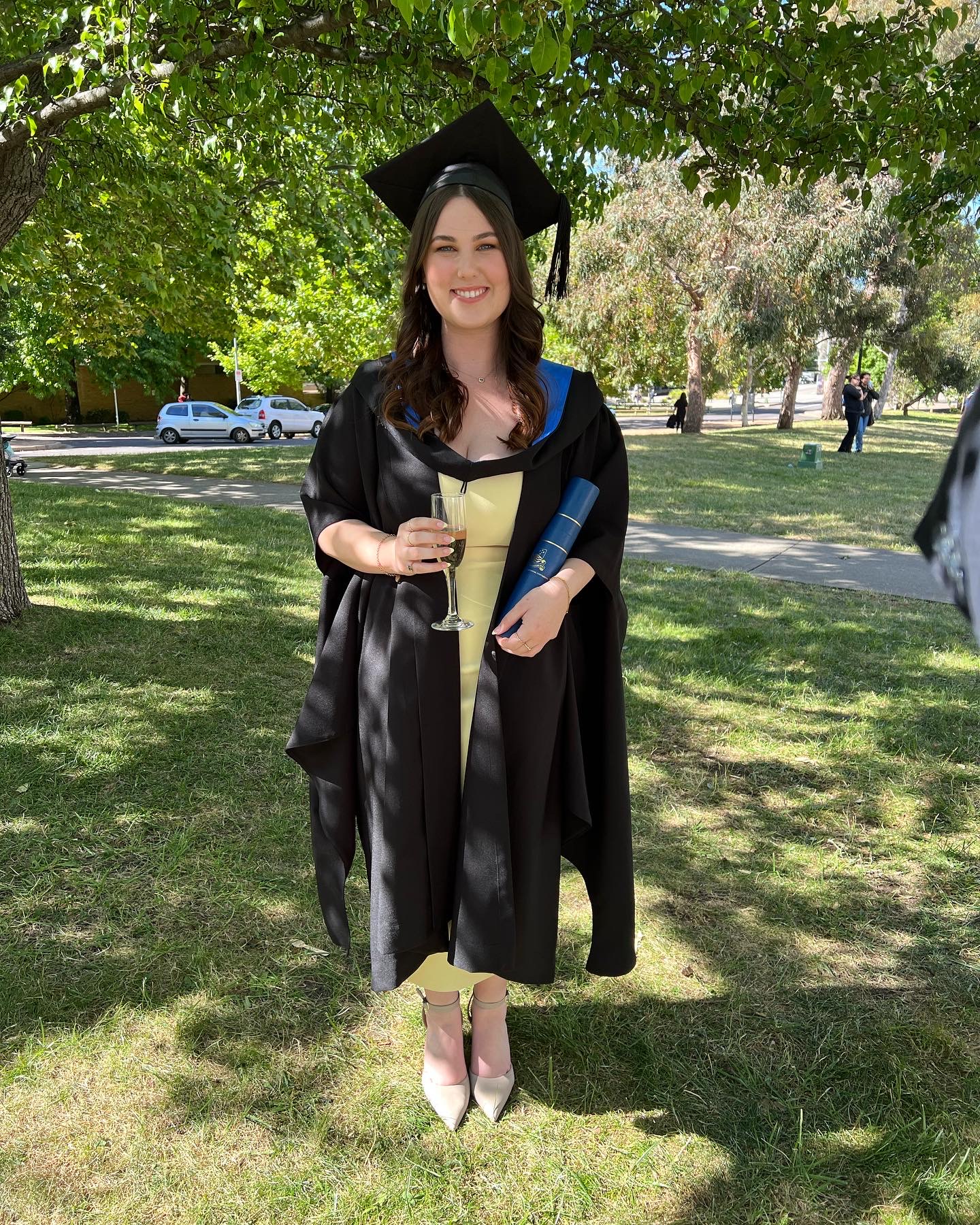 Sophie smiles in her graduation cap and gown, holding her diploma outdoors underneath a tree.
